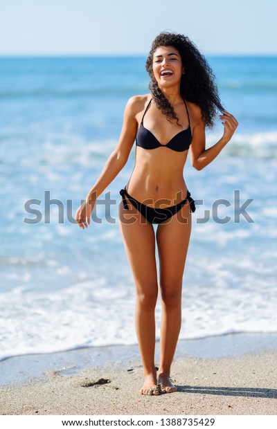熱帯の海岸で微笑む 美しい体を持つ若いアラビア人女性の水着 黒いビキニを着た巻き毛の女性 の写真素材 今すぐ編集