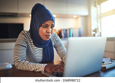 Young Arabic Woman Wearing A Hijab Sitting At Her Kitchen Table Using A Laptop While Working From Home