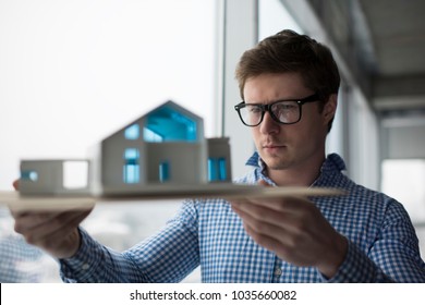Young architect holding a home model in loft modern office. Architectural project concept. University student wearing stylish hipster glasses planning engineering design, brainstorming at workplace - Powered by Shutterstock