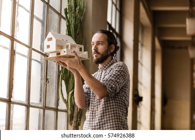 Young architect is looking on the new model in the old industrial space with big factory windows. Man is standing in front of window and holding small model of the house. Color toned image.