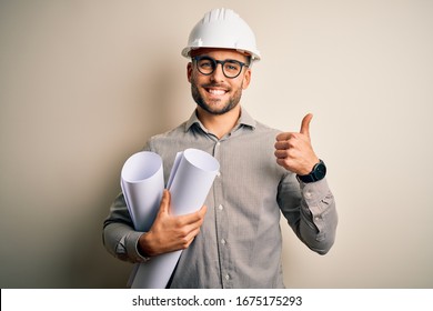 Young architect man wearing contractor helmet holding project paper plan over isolated background happy with big smile doing ok sign, thumb up with fingers, excellent sign