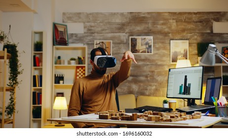 Young architect using virtual reality goggles modeling buildings in home office.
