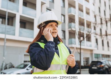 Young architect woman in white hardhat and safety vest with digital tablet talking on smartphone near modern building. Female engineer. Logistics, shipping and construction worker on the phone - Powered by Shutterstock