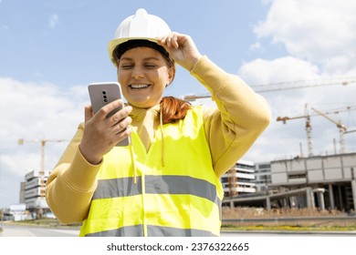 Young Architect woman in white hardhat and safety vest with digital tablet using smartphone outdoors. Female engineer. Logistics and construction worker near modern building. Engineering application - Powered by Shutterstock