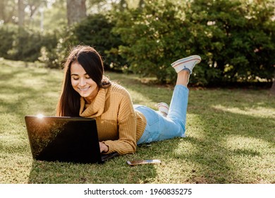 A young Argentinian female student in a yellow sweater lying on the grass and using a laptop