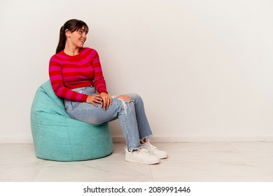 Young Argentinian woman sitting on a puff isolated on white background looks aside smiling, cheerful and pleasant.