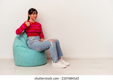 Young Argentinian woman sitting on a puff isolated on white background person pointing by hand to a shirt copy space, proud and confident