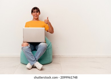 Young Argentinian woman working with pc sitting on a puff isolated on white background smiling and raising thumb up