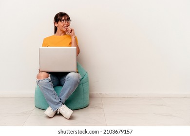 Young Argentinian woman working with pc sitting on a puff isolated on white background relaxed thinking about something looking at a copy space.