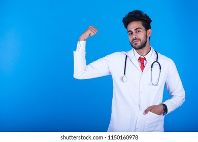 Young Arrogant Doctor Put Hands In The Pocket Of The Coat Raising One Arm Showing His Biceps On A Blue Background.