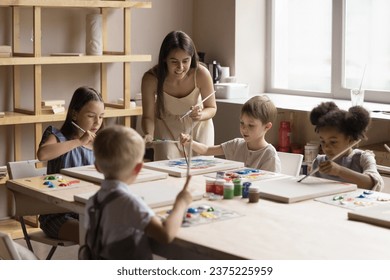 Young artist female teacher lead art-class for little children, multi-ethnic kids drawing pictures with paints on canvas seated at table in art-studio. Talent development, creative hobby, education - Powered by Shutterstock