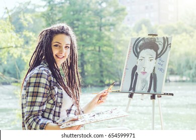 Young Artist Girl Painting A Self Portrait In A Park Near Lake - Painter Woman With Dreadlocks Hairstyle Working On Her Art In The City Outdoor - Concept Of People Expressing Arts 