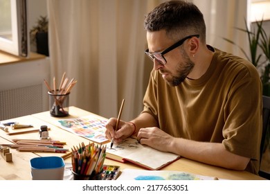 A young artist makes a brush sketch at his desk. Portrait of a creative person in his studio. Creative studio, lifestyle, the process of creating a work of art, the search for inspiration. - Shutterstock ID 2177653051