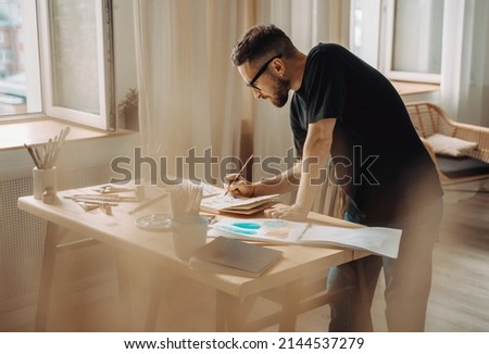 A young artist makes a sketch with a brush in the workshop. A creative person is standing over a desktop with a brush in his hand. Creative studio, lifestyle, the process of creating a work of art.
