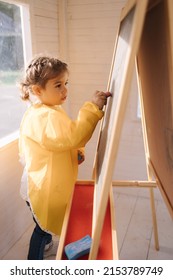 Young artist paints on wooden boards with chalk. Cute girl in yeallow raincoat