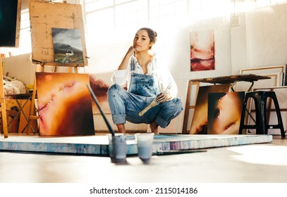 Young artist squatting on the floor while working in her art studio. Female painter looking at the camera and holding a paintbrush. Creative young woman making a new painting on a canvas.