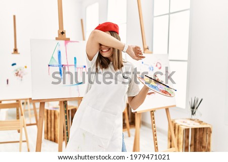 Young artist woman painting on a canvas at art studio smiling cheerful playing peek a boo with hands showing face. surprised and exited 