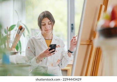 Young artist working in the art studio, she is connecting online with her smartphone and chatting - Powered by Shutterstock