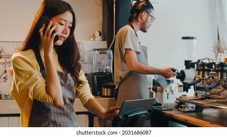 Young Asia female barista taking order by mobile phone and digital tablet standing behind bar counter while talking with customer on mobile phone at cafe restaurant. Owner small business concept. - Powered by Shutterstock