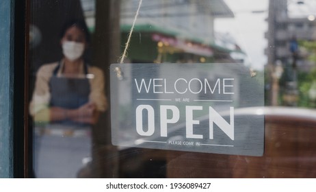 Young Asia girl wear face mask turning a sign from closed to open sign on door looking outside waiting for clients after lockdown. Owner small business, food and drink, business reopen again concept.