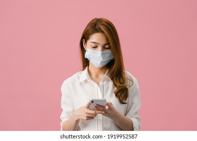 Young Asia girl wearing medical face mask using mobile phone with dressed in casual clothing isolated on pink background. Self-isolation, social distancing, quarantine for corona virus prevention. - Powered by Shutterstock