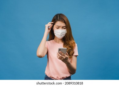 Young Asia girl wearing medical face mask using mobile phone with dressed in casual clothing isolated on blue background. Self-isolation, social distancing, quarantine for corona virus prevention. - Powered by Shutterstock