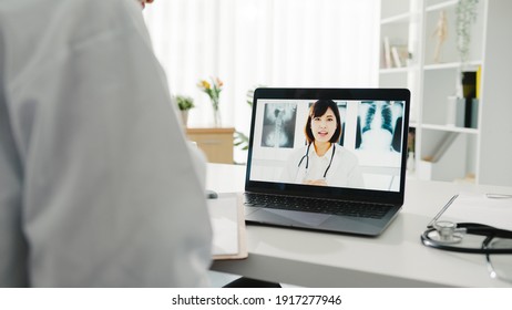Young Asia Lady Doctor In White Medical Uniform Using Laptop Talking Video Conference Call With Senior Doctor At Desk In Health Clinic Or Hospital. Social Distancing, Quarantine For Corona Virus.