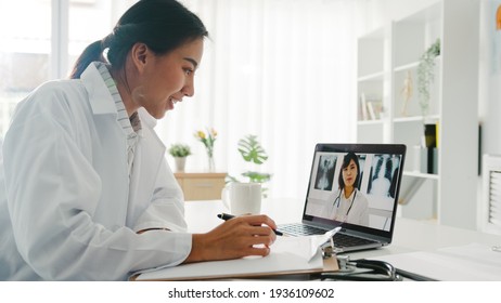Young Asia Lady Doctor In White Medical Uniform Using Laptop Talking Video Conference Call With Senior Doctor At Desk In Health Clinic Or Hospital. Social Distancing, Quarantine For Corona Virus.