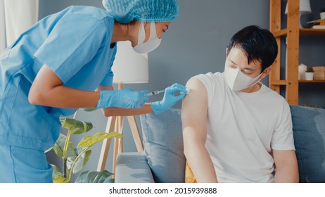 Young Asia Lady Nurse Giving Covid-19 Or Flu Antivirus Vaccine Shot To Male Patient Wear Face Mask Protection From Virus Disease Sit On Couch In Living Room At House. Vaccination Concept.