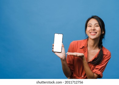 Die junge Asienfrau zeigt einen leeren Smartphone-Bildschirm mit positivem Ausdruck, lächelt im Allgemeinen, gekleidet in lockerer Kleidung und fühlt sich glücklich auf blauem Hintergrund. Handy mit weißem Bildschirm in weiblicher Hand.