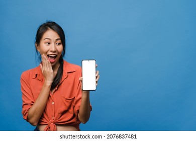 Young Asia Lady Show Empty Smartphone Screen With Positive Expression, Smiles Broadly, Dressed In Casual Clothing Feeling Happiness On Blue Background. Mobile Phone With White Screen In Female Hand.