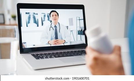 Young Asia Lady Using Computer Laptop Talk About A Disease In Video Conference Call With Senior Doctor Online Consultation In Living Room At Home. Social Distancing, Quarantine For Coronavirus Concept