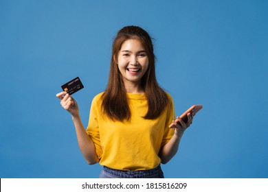 Young Asia lady using mobile phone and credit bank card with positive expression, dressed in casual cloth and looking at camera isolated on blue background. Happy adorable glad woman rejoices success.