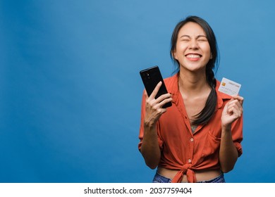 Young Asia lady using phone and credit bank card with positive expression, smiles broadly, dressed in casual clothing and stand isolated on blue background. Happy adorable glad woman rejoices success.