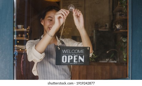 Young Asia Manager Girl Changing A Sign From Closed To Open Sign On Door Cafe Looking Outside Waiting For Clients After Lockdown. Owner Small Business, Food And Drink, Business Reopen Again Concept.