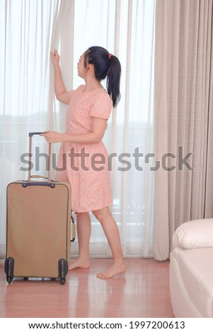 Young Asia woman traveler in pink dress with her luggage arrives at the hotel room and open curtain for enjoying an outside view, Happy women lifestyle with holiday summer travel vacation concept