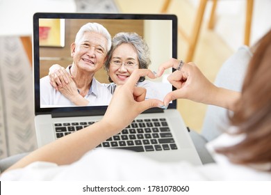 young asian adult daughter staying at home forming a heart shape with hands while talking to senior parents online via video chat using laptop computer