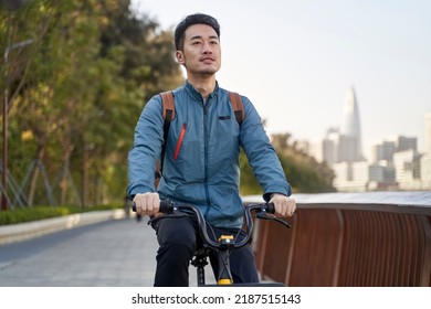 Young Asian Adult Man Riding A Bike In Riverfront Park With City Skyline In Background