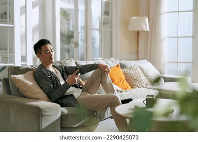 young asian adult man sitting on family couch in living room at home looking at cellphone - Powered by Shutterstock