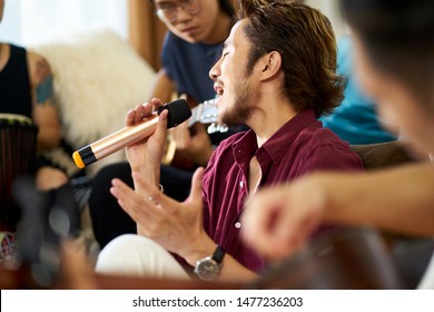 Young Asian Adult Men Rock Band Members Rehearsing Singing Playing Musical Instruments In Living Room Of A House