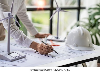 Young Asian architect with blueprints working on lapotp in office. Career working hard on wooden table, painting with windmills, designing model of renewable wind house building. and sunlight - Powered by Shutterstock