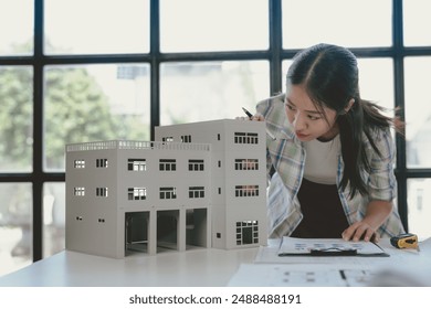 Young asian architect carefully reviews blueprints and uses a scale model to plan a new office building in the city - Powered by Shutterstock