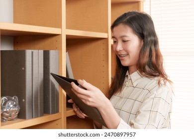 Young Asian architect organizing files in office. Woman wearing plaid shirt handling documents on shelf, conveying organization and efficiency in professional workplace environment. - Powered by Shutterstock