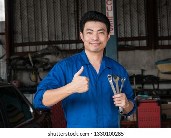 Young Asian Auto Mechanic In Uniform Holding Wrenches And Showing Thumbs Up In The Garage. Repair, Car Service And Maintenance Concept.