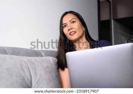Young asian Bashkir woman works on computer at home, sitting on the sofa, smiling