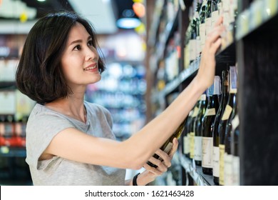 Young Asian Beautiful Casual Cloth Woman Holding Shopping In Supermarket. She Is Choosing Nice Red Wine Bottle Picking Up From Alcohol Shelf At Wine Store Shop While She Looking At Product And Smile.