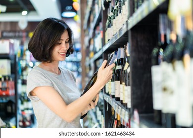 Young Asian Beautiful Casual Cloth Woman Holding Shopping In Supermarket. She Is Choosing Nice Red Wine Bottle Picking Up From Alcohol Shelf At Wine Store Shop While She Looking At Product And Smile.