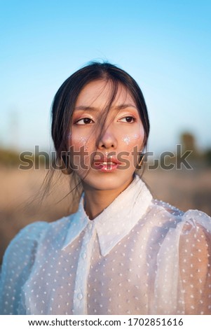Young asian young beautiful model woman with black hair in a white transparent shirt from Zara. close-up portrait in the field, glitter makeup, and smokey eyes—capturing modern fashion allure.