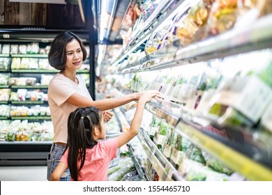 Young Asian beautiful mother holding grocery basket with her child walking in supermarket. She is choosing green salad vegetable picking up from shelf with her little daughter. Shopping for healthy. - Powered by Shutterstock