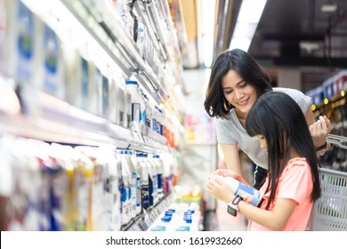 Young Asian beautiful mother holding grocery basket with her child walking in supermarket. She is choosing daily milk product picking up from shelf with her little daughter. Shopping for healthy.